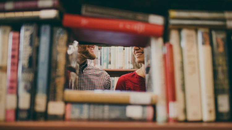 Man and woman smiling at each other behind a shelf of books