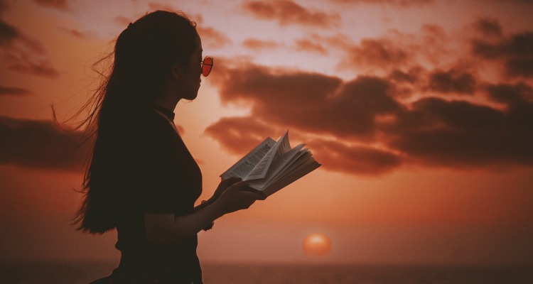 Silhouette of a girl with sunglasses reading a book at sunset with a red-orange sky in the background