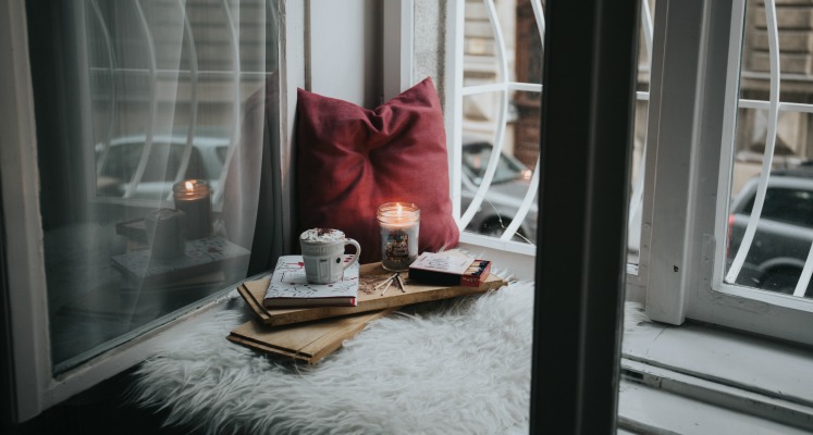 Cozy window seat reading nook with books, cushion, candle, and hot cocoa