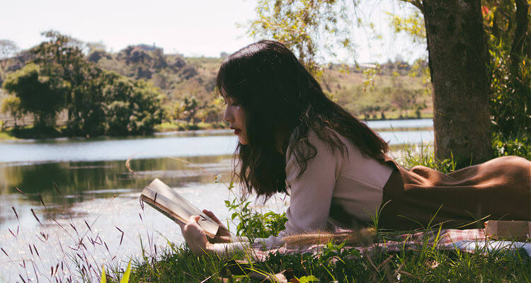 Woman lying on the grass reading a book by a pond