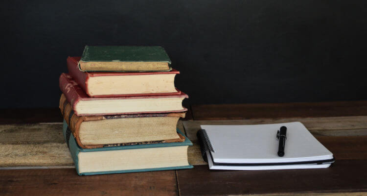 Stack of books on a wooden table, with open notebook and pen