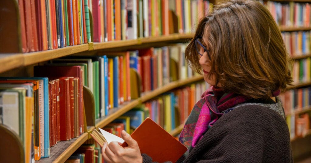 Middle-age woman with a colorful pink scarf and glasses browsing a book in the library.