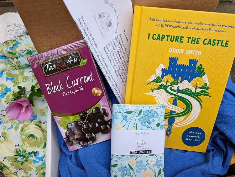 Summer reading subscription box featuring I Capture the Castle novel, box of black currant tea, and floral fabric tea wallet.